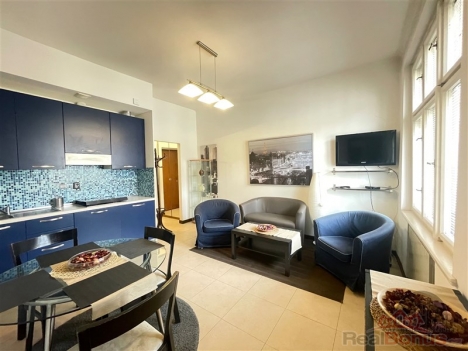 Kuchy�sk� kout s tmav� modr�mi sk���kami, sv�tl� dla�ba a modern� osv�tlen�, vedle ob�vac� z�ny s pohovkami a velk�mi okny.