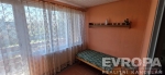 Mal� pokoj s velk�mi okny, z�clony propou�t�j�c� sv�tlo a dekorativn� poli�ka. Kobercov� podlaha a �tuln� styl.